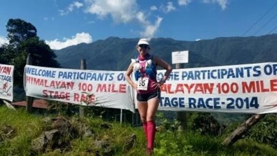 A woman stands beside a banner that reads "WELCOME PARTICIPANTS OF HIMALAYAN 100 MILE STAGE RACE - 2014."